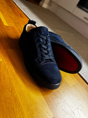Mörkblå mockasneakers från Louboutin - Snygga mörkblå sneakers i mocka från Christian Louboutin med klassisk röd sula och diskret logga på tungan. Skorna har rund tå, platt sula och snörning. Insidan är i ljust skinn och sulan har den ikoniska Louboutin-signaturen.