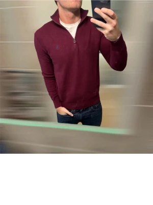 Vinröd stickad tröja Polo Ralph Lauren - Säljer en vinröd stickad tröja från Polo Ralph Lauren med half zip och hög krage. Tröjan har lång ärm och den klassiska blå loggan broderad på bröstet. Perfekt till jeans för en clean och stilren look.