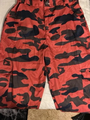 Röda cargopants med camomönster - Säljer ett par riktigt feta cargopants i rött med svart och mörkgrå camouflagemönster. Byxorna har stora sidofickor och klassisk cargostil. Perfekta för dig som vill sticka ut och gillar streetwear-vibe. Materialet känns slitstarkt och de har bälteshällor i midjan.