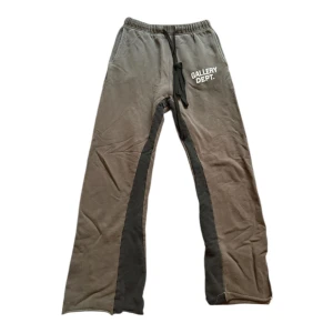 Mjukisbyxor från Gallery Dept  - Grå mjukisbyxor från Gallery Dept. med svarta och beige paneler längs benen. Byxorna har elastisk midja med snörning och tryckt logga framtill. Raka ben med slitsar bak vid benslut. Perfekta för en chill och avslappnad stil.