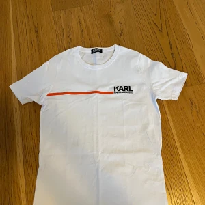 Vit t-shirt från Karl Lagerfeld - Snygg vit t-shirt från Karl Lagerfeld med en orange linje över bröstet och logga tryckt på vänster sida. Klassisk rund hals och korta ärmar. Tillverkad i mjuk bomull som känns skön mot huden. Perfekt för en clean och stilren look.