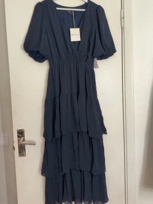 Marinblå långklänning från Willa - Säljer en otroligt fin midiklänning i mörkblå (navy) färg. Klänningen har en smickrande omlottliv med V-ringning, markerad midja och vackra lager i kjolen som ger ett fint fall. Puffärmar som ger en feminin och elegant look. Perfekt till både vardag, fest, bröllop, student, dop eller middagar 🌙 Lätt och luftig – väldigt bekväm att bära. 👗 Storlek: EU 38 🎨 Färg: Navy blue / mörkblå 📏 Längd: Midi ✨ Skick: helt ny med prislapp. Köpt för 1399kr