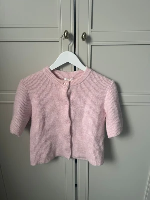 Ljusrosa stickad kofta från Gina Tricot - Supersöt ljusrosa stickad kofta från Gina Tricot med korta ärmar. Koftan har ribbad struktur och knäpps med matchande knappar framtill. 