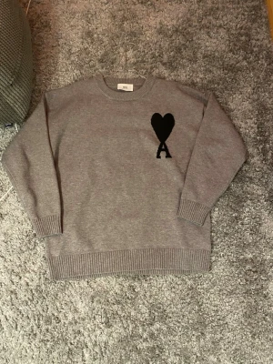 Grå stickad tröja från Ami - Säljer en grå stickad tröja från Ami  med svart hjärta och bokstaven A broderat på bröstet. Tröjan har rund hals, ribbade muddar och en relaxed passform. Perfekt till jeans för en clean look. Passar som M och L 🤟🏽🤟🏽