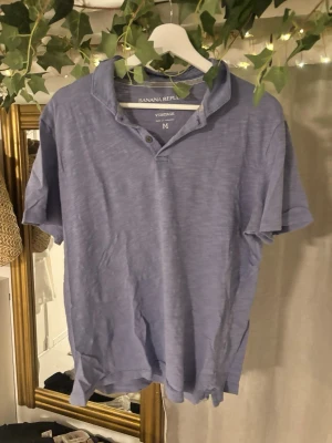 Blå kortärmad t-shirt Banana Republic - Säljer en blå kortärmad t-shirt från Banana Republic Vintage. T-shirten har en klassisk krage med knappdetalj och diskreta ränder på insidan av kragen. Tillverkad i mjuk bomull för en skön känsla.