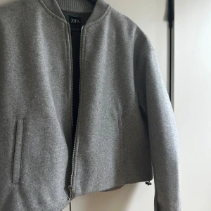 Grå bomberjacka från Zara  - Snygg grå bomberjacka från Zara i ullmix med klassisk ribbad krage och raka fickor. Jackan har en rak, något croppad passform och stängs med silverfärgad dragkedja. Perfekt för dig som gillar stilren och enkel design.