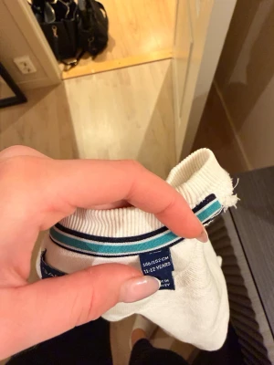 Vit sweatshirt från Gant med tryck - Vit långärmad sweatshirt från Gant med rund hals och blått tryck på bröstet med texten 'Championships Båstad 2018 Sweden'. Kragen har en blå och turkos randig detalj. Perfekt för en sportig och avslappnad stil.