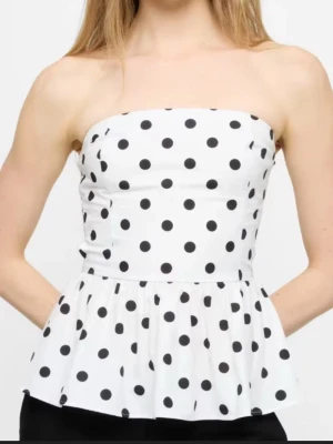 Prickig vit tubtopp med volang - Säljer en söt axelbandslös tubtopp i vitt med stora svarta prickar. Toppen har en volang nedtill som ger en lekfull look och är perfekt för varma dagar. Passar dig som gillar trendiga och feminina plagg. Nypris: 500