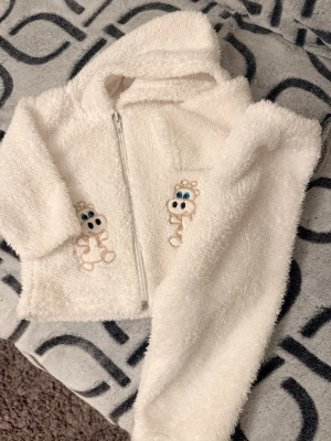 Vit fluffig väst med dragkedja - Supersöt vit fluffig väst med huva och dragkedja framtill. Västen har ett broderat motiv av en gullig figur med blå ögon på bröstet. Perfekt för dig som gillar mysiga och lekfulla plagg.