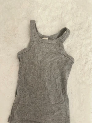 Grå ribbad linnetopp  - Säljer en enkel och stilren grå ribbad linnetopp från H&M Basics. Toppen är ärmlös med breda axelband och har en tight passform. Perfekt att matcha med jeans eller kjol för en clean look.