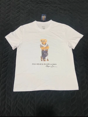 Vit Polo Bear t-shirt från Ralph Lauren - Vit t-shirt från Ralph Lauren med ikoniskt Polo Bear-tryck på bröstet. Klassisk rund hals och korta ärmar. Slim fit-modell i mjuk bomull, perfekt för en clean och stilren look.