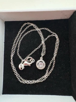 Silverhalsband med gnistrande cirkelhänge 925 Stämpel på - Säljer ett stilrent halsband i äkta silver med ett runt hänge prytt av en glittrande sten i mitten och små stenar runtom. Kedjan är tunn och smidig med karbinlås. Perfekt för dig som gillar klassiska smycken med lite extra bling.