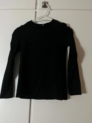 Svart ribbad topp från Lindex Kids - En svart långärmad topp från Lindex Kids i storlek 134/140. Toppen har ribbad struktur och puffade axlar som ger en trendig look. Tillverkad i mjukt material som känns skönt mot huden. Perfekt till jeans eller kjol för en enkel men snygg outfit.