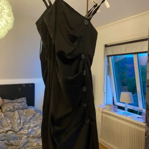 Svart kort klänning SisterS Point XS - Svart kort klänning från SisterS Point med smala axelband och stora dekorativa knappar framtill. Snygg drapering längs sidan och figurnära passform. Perfekt för dig som gillar stilrena och trendiga plagg.