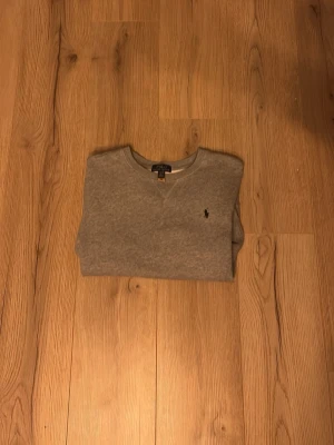 Grå sweatshirt från Polo Ralph Lauren - Klassisk grå sweatshirt från Polo Ralph Lauren med rund halsringning och broderad marinblå logga på bröstet. Tröjan har ribbade muddar vid ärmslut och nederkant, samt diskreta sömdetaljer vid halsen. Perfekt för en chill och stilren look.