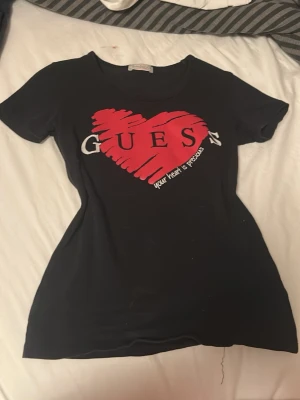 Svart t-shirt med rött hjärta Guess - Svart t-shirt från Guess Jeans med ett stort rött hjärta och texten 'Guess' samt 'your heart is precious' på bröstet. Klassisk rund halsringning och korta ärmar. T-shirten har en figurnära passform och är tillverkad i mjukt material.