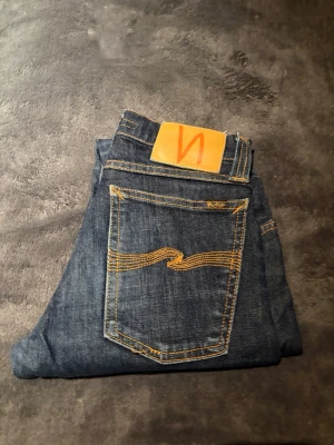  Nudie Jeans - Säljer ett par mörkblå skinny jeans från Nudie Jeans med klassisk orange söm och Nudie-logga på bakfickan. Jeansen har fem fickor, knappgylf och är tillverkade i slitstarkt jeansmaterial. Perfekta för dig som gillar en smal passform och tidlös stil.