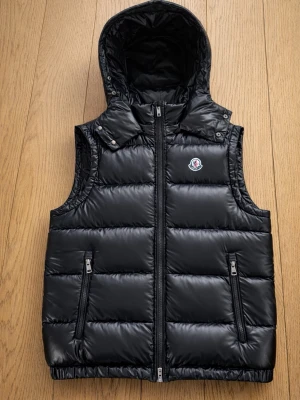 -Moncler Väst svart- - Säljer en äkta Moncler väst i mycket fint skick. Västen är superbekväm, lätt att bära och samtidigt riktigt varm – perfekt för vår, höst eller kyliga sommarkvällar.  Äkta Moncler Av- och påtagbar luva Skön passform och varm Stilren och tidlös modell  Det finns 2 små hål på västen, inget som stör funktionen men därför är priset satt därefter.