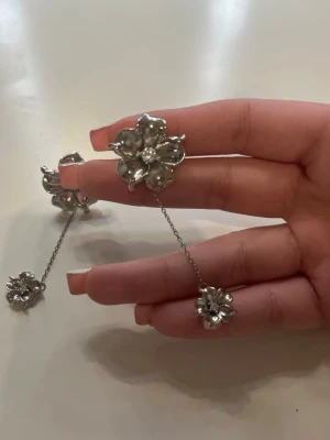 Silverfärgade blomörhängen med kedja - Säljer ett par statement örhängen i silverfärg med stora blommor och gnistrande sten i mitten. Örhängena har en tunn kedja som binder ihop två blommor för en unik look. Perfekta för att lyfta vilken outfit som helst. De är ifrån LYLA och kostar i vanliga fall 279 kr. (ALDRIG ANVÄNDA!)