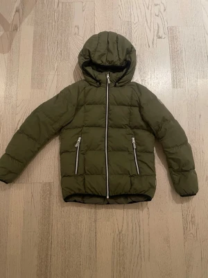 Grön dunjacka från Reima - Säljer en grön dunjacka från Reima med huva och två fickor med dragkedja. Jackan har quiltad design och vita detaljer på dragkedjorna. Perfekt för kalla dagar och har en snygg, enkel look som funkar till det mesta.