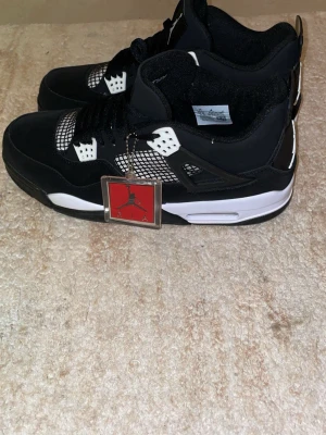 Nike Air Jordan 4 svart/vit - Säljer ett par Nike Air Jordan 4 i svart och vitt med klassisk meshpanel och detaljer i plast. Skorna har snörning, platt sula och den ikoniska Jumpman-taggen. Perfekta för dig som vill ha en stilren och sportig sneaker med retrovibbar.