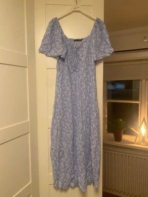 Blommig blå klänning från lager 157 - Supersöt långklänning från lager 157 i ljusblått med vitt blommönster. Klänningen har korta ärmar och en liten snörning framtill. Perfekt till midsommar