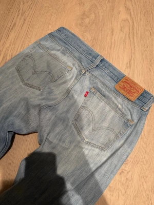 vintage Levi's 501  - Ljusblå Levi's 501 jeans med klassisk femficksdesign och knappgylf. Jeansen har en rak passform och är tillverkade i jeansmaterial. Snygga slitningar och hål på vänster lår ger en cool, avslappnad vibe. Levi's patch och röd etikett bak på midjan.