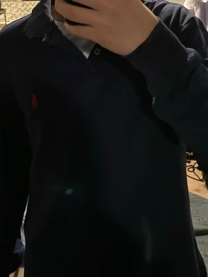 Marinblå pikétröja från Polo Ralph Lauren - Klassisk marinblå pikétröja från Polo Ralph Lauren i storlek S. Tröjan har lång ärm, krage och den ikoniska röda logotypen broderad på bröstet. Perfekt till jeans eller chinos för en clean och tidlös look.