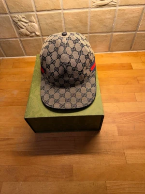 Gg beige GG-mönstrad keps M - Säljer en beige keps från Gg  med klassiskt GG-mönster i mörkblått och röda/blå ränder på sidorna. Justerbar rem bak i skinn och insida i svart tyg. Perfekt accessoar för dig som vill ha en ikonisk och stilren look.