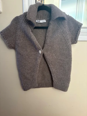 Beige stickad kofta från Zara - Kortärmad beige stickad kofta från Zara med bred krage och en knapp framtill. Koftan har ribbade muddar och är perfekt för lager på lager under höst och vår. Snygg att styla över en t-shirt eller topp för en chill look.