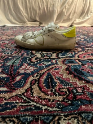 Philippe Model  - Säljer ett par vita Philippe Model sneakers med grå detaljer och en cool gul häl. Skorna har snörning med logotyp på skosnörena och en klassisk låg profil. Tillverkade i skinn och mocka, med svart sula och rund tå.