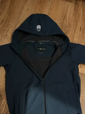 Mörkblå vindjacka från Sail Racing - Säljer en mörkblå vindjacka från Sail Racing med huva och hel dragkedja. Inga skador.                 Jackan har diskreta loggor på bröstet och ärmen, samt två fickor framtill. Materialet är slitstark polyester och insidan är mjukt fodrad för extra komfort. Perfekt för blåsiga dagar.