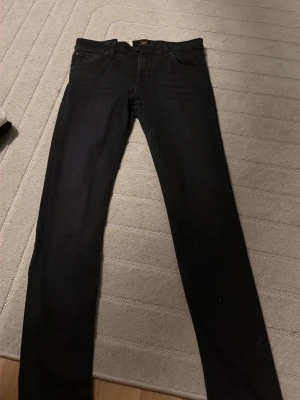 Svarta skinny jeans från Lee - Säljer ett par svarta skinny jeans från Lee med klassisk femficksdesign och smal passform. Jeansen har normal midja och är tillverkade i stretchig bomullsblandning för extra komfort. Perfekta till sneakers eller boots.
