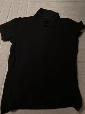 Svart pikétröja med krage - En klassisk svart pikétröja med korta ärmar och krage. Tröjan har knappar framtill vid halsen och är tillverkad i ett mjukt och bekvämt material. Perfekt till jeans eller shorts för en enkel och stilren look.