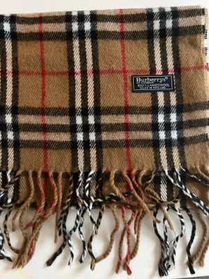 Rutig halsduk från Burberry - Klassisk halsduk från Burberrys i 100% lammull med ikoniskt rutigt mönster i beige, svart, vitt och rött. Halsduken har fransar nedtill och en mjuk, varm känsla. Perfekt för dig som vill ha en snygg accessoar. 