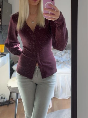 Waist focus cardigan från Nelly - Säljer en vinröd blus med v-ringning och långa ärmar. Blusen har en figurnära passform och knäppning framtill. Perfekt att styla med jeans för en snygg look. Materialet känns mjukt och följsamt. Är bara använd någon gång och är i nyskick. Storlek Xxs