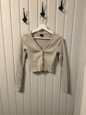 Beige ribbad kofta från Gina Tricot - En beige ribbad kofta från Gina Tricot i storlek S. Koftan är croppad med v-ringning, långa ärmar och knappar framtill.🤩 Perfekt att slänga över ett linne eller t-shirt för en chill look.🥰Går jättebra att använda "Köp nu"! Vi skickar inom 1-2 dagar💕 Bara att skriva vid funderingar🤗