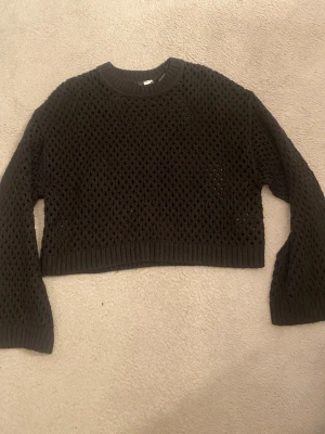 Svart tröja - Säljer en svart croppad stickad tröja med hålmönster och långa ärmar. Tröjan har en ribbad rund halsringning och muddar vid ärmslut och nederkant. Perfekt att styla över ett linne eller t-shirt för en cool lager-på-lager-look.