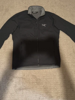 Svart vindjacka från Arc'teryx - Säljer en svart vindjacka från Arc'teryx med grå insida och hög krage. Jackan har dragkedja framtill, två sidofickor med dragkedja och en bröstficka. Materialet är lätt och smidigt, perfekt för blåsiga dagar. Diskret Arc'teryx-logga på bröstet.