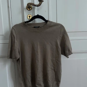 Beige/brun stickad kortärmad topp  - Stickad kortärmad topp i mjuk kvalitet från Lindex. Toppen är i använt skick. Det finns en liten defekt baktill (se närbild), vilket återspeglas i priset. I övrigt hel och fullt användbar.