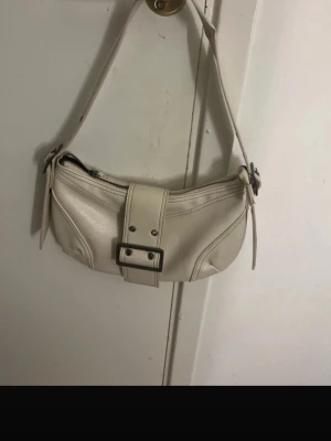 Beige axelväska med spänne - Säljer en beige axelväska i skinnimitation med coolt spänne framtill och dragkedja upptill. Väskan har en bred axelrem och stilrena sömmar som ger en snygg look. Perfekt storlek för det viktigaste och passar till många outfits.