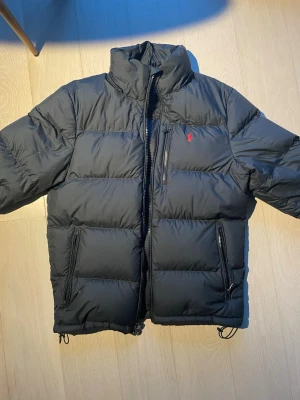 Svart pufferjacka från Polo Ralph Lauren - Nypris är 5500kr. Har kvitto. Säljer en svart pufferjacka från Polo Ralph Lauren med hög krage och dragkedja. Jackan har tre fickor med dragkedja och den klassiska röda loggan på bröstet. Perfekt för kalla dagar och riktigt snygg till streetwear-stilen.