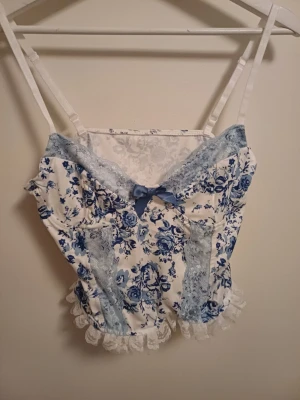 Blommig spetstopp med rosett - Supersöt topp med blått och vitt blommönster, spetsdetaljer i ljusblått och vit volangkant. V-ringad med liten blå rosett framtill och justerbara axelband. Perfekt för sommaren. Jag har aldrig använt den 