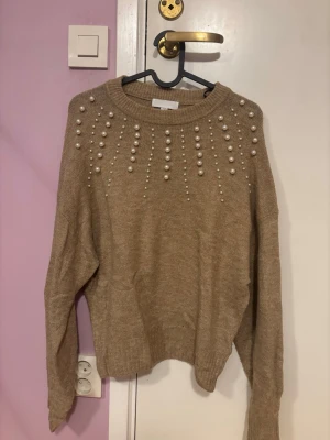 Beige stickad tröja med pärlor H&M - Säljer en beige stickad tröja från H&M i storlek XS. Tröjan har långa ärmar och är dekorerad med pärlor över axlar och bröst, vilket ger en snygg och trendig look. Perfekt för dig som vill ha något extra till hösten eller vintern.