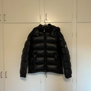  Moncler Maya Jacka - Säljer en svart Maya jacka från Moncler med glansig finish, huva och dragkedja framtill. Jackan har två fickor med dragkedja, Moncler-logga på ärmen och quiltad design. Perfekt för kalla dagar och riktigt snygg streetstil. NFC finns samt fungerar. Använd ca 1 vinter, så väldigt bra skick. Säljs då den tyvärr inte passar längre. Vid funderingar eller liknande är det bara att höra av sig!