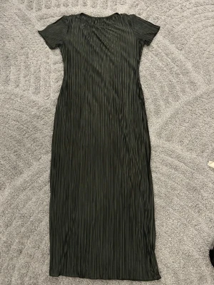 Mörkgrön  långklänning - Säljer en långklänning i mörkgrönt med plisserad struktur och korta ärmar. Klänningen har rund halsringning och ett snyggt, glansigt tyg som ger en elegant känsla. Perfekt för dig som gillar stilrena och moderna plagg.