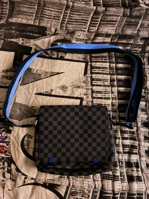 Louis Vuitton rutig axelväska - Snygg axelväska från Louis Vuitton i svart och grått med klassiskt rutigt mönster. Väskan har en bred blå axelrem och detaljer i blått samt präglad logotyp. Tillverkad i tåligt skinnmaterial och perfekt för dig som vill ha en stilren och modern look. Inget kvitto eller dustbag 