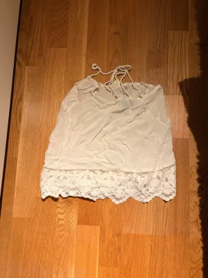 Vit linnetopp med spetskant - Säljer en söt vit linnetopp med tunna axelband och dekorativ spetskant längst ner. Toppen har en luftig passform och är perfekt för varma dagar. Materialet känns lätt och toppen knyts i nacken för en enkel och stilren look.