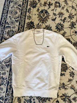 Lacoste vit tröja  - Säljer en clean vit sweatshirt från Lacoste med klassisk rund hals och broderad krokodil-logga på bröstet. Tröjan är i mjuk bomull och har långa ärmar. Perfekt för en chill och stilren look.