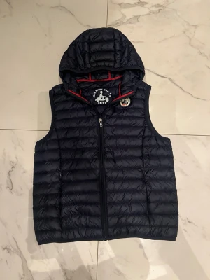Marinblå Väst - Marinblå dunväst från JOTT med luva. Skön passform, storlek 12/14 (160 - 165 cm). Den kommer med en bärbar förvaringspåse och har två zip fickor. Pris kan diskuteras!